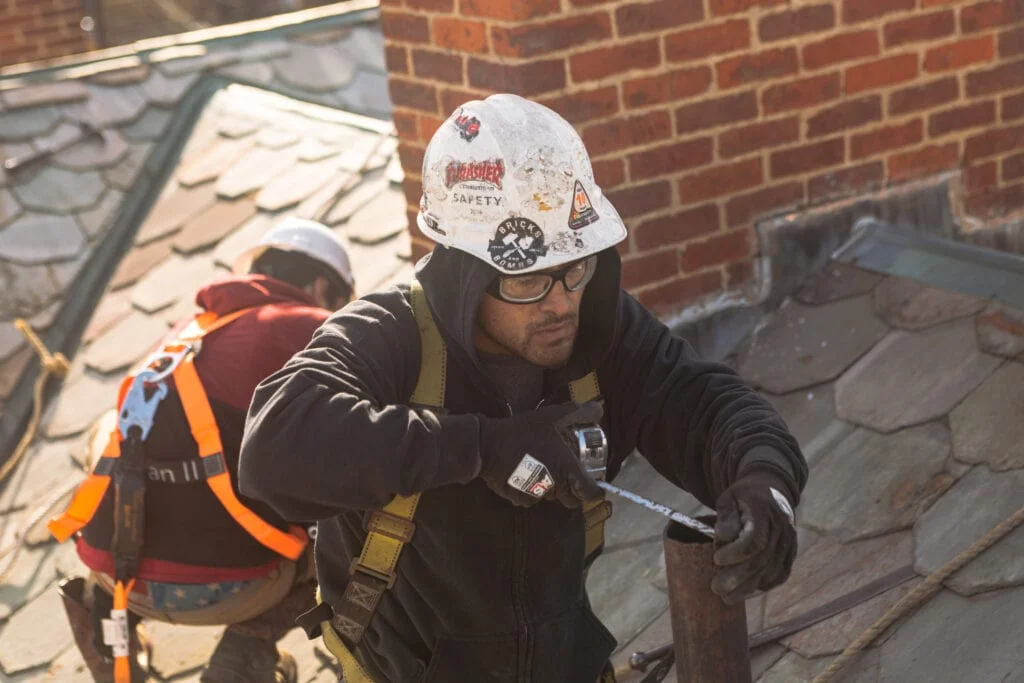 paul measuring a vent pipe in front of a chimney while kneeling on a slate roof